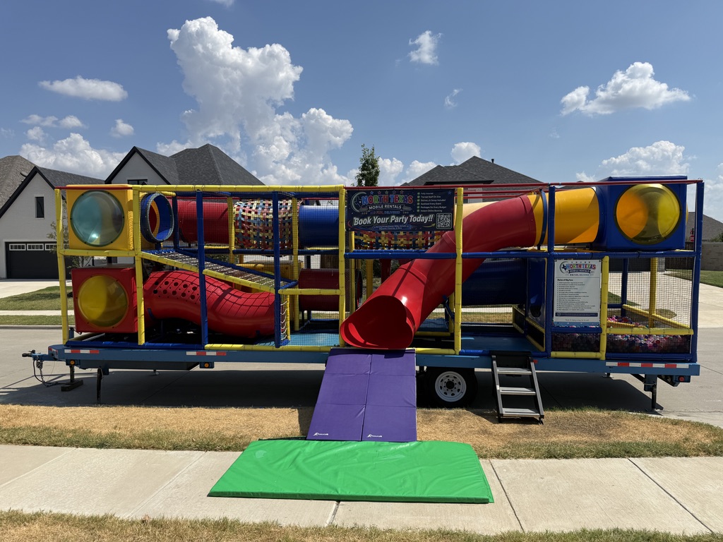 Side view of the Signature Mobile Playground trailer with red tube slide, tunnels, and ball pit, parked at the curb on a North Texas street